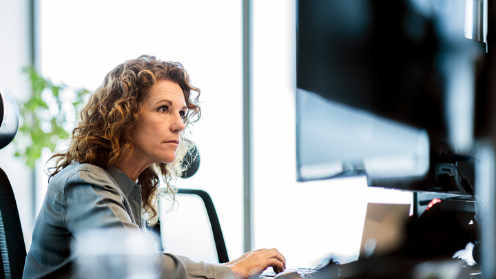 Professional woman with curly hair working at a desktop computer in a modern office, focused on the screen while typing, with natural light and a blurred background.