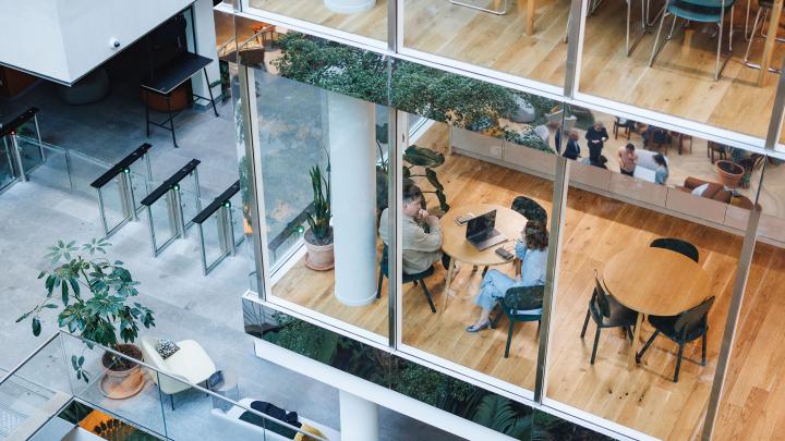 Modern office atrium with glass meeting rooms, wooden floors, security turnstiles, indoor plants, and two people in discussion at a round table.