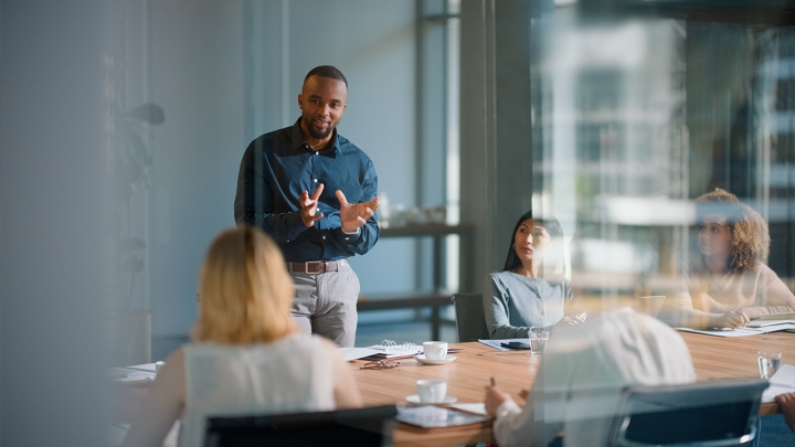 Professional leading a meeting in a modern glass-walled boardroom, speaking to seated colleagues around a wooden conference table with notebooks, coffee cups and water glasses visible.