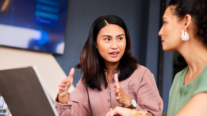 Two colleagues in a modern office discussing a project, with one woman gesturing as she explains something while the other listens; a laptop is open on the table beside them.