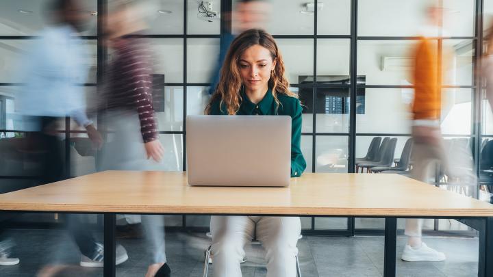 Person working on a laptop in a modern office while colleagues move around in the background.