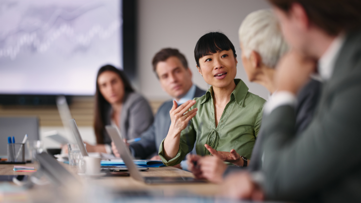  A diverse group of business professionals participating in a meeting around a conference table, with a woman in a green blouse leading the discussion as colleagues listen. Laptops and documents are visible on the table.
