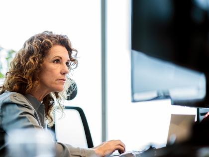 Professional woman with curly hair working at a desktop computer in a modern office, focused on the screen while typing, with natural light and a blurred background.