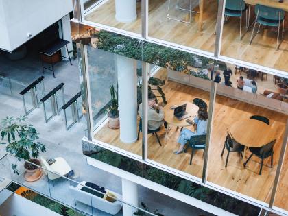 Modern office atrium with glass meeting rooms, wooden floors, security turnstiles, indoor plants, and two people in discussion at a round table.