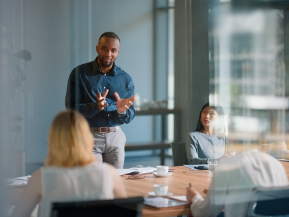 Professional leading a meeting in a modern glass-walled boardroom, speaking to seated colleagues around a wooden conference table with notebooks, coffee cups and water glasses visible.