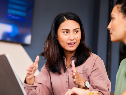 Two colleagues in a modern office discussing a project, with one woman gesturing as she explains something while the other listens; a laptop is open on the table beside them.
