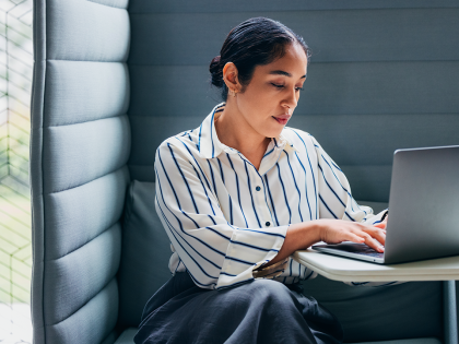 Business professional working on a laptop in a private office booth, wearing a striped shirt and seated at a small desk in a modern workspace with natural light from nearby windows.