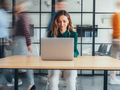 Person working on a laptop in a modern office while colleagues move around in the background.