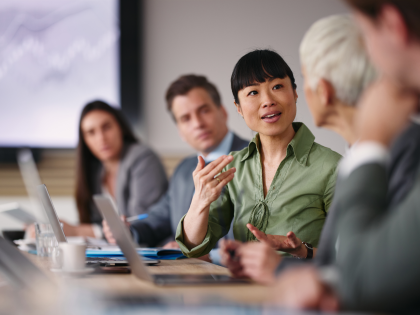  A diverse group of business professionals participating in a meeting around a conference table, with a woman in a green blouse leading the discussion as colleagues listen. Laptops and documents are visible on the table.