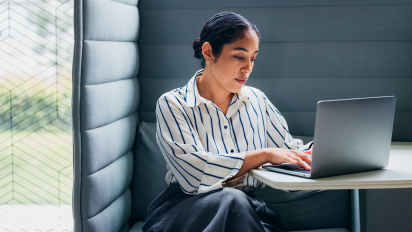 Business professional working on a laptop in a private office booth, wearing a striped shirt and seated at a small desk in a modern workspace with natural light from nearby windows.